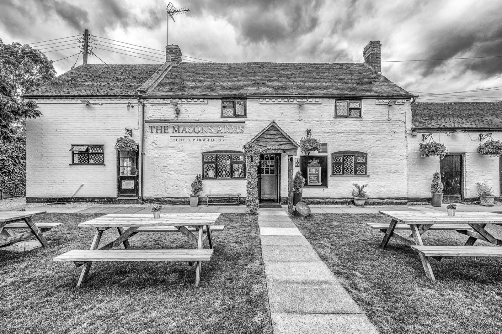 Black-and-white photo of The Masons Arms' whitewashed, half-timbered exterior with 'The Masons Arms - Country Pub & Dining' signage and picnic tables on the front lawn.